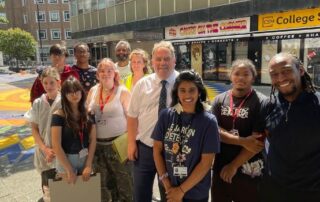Mayor Perry stands with students and artists at the new Croydon Compass artwork in Croydon Square. It is very sunny.